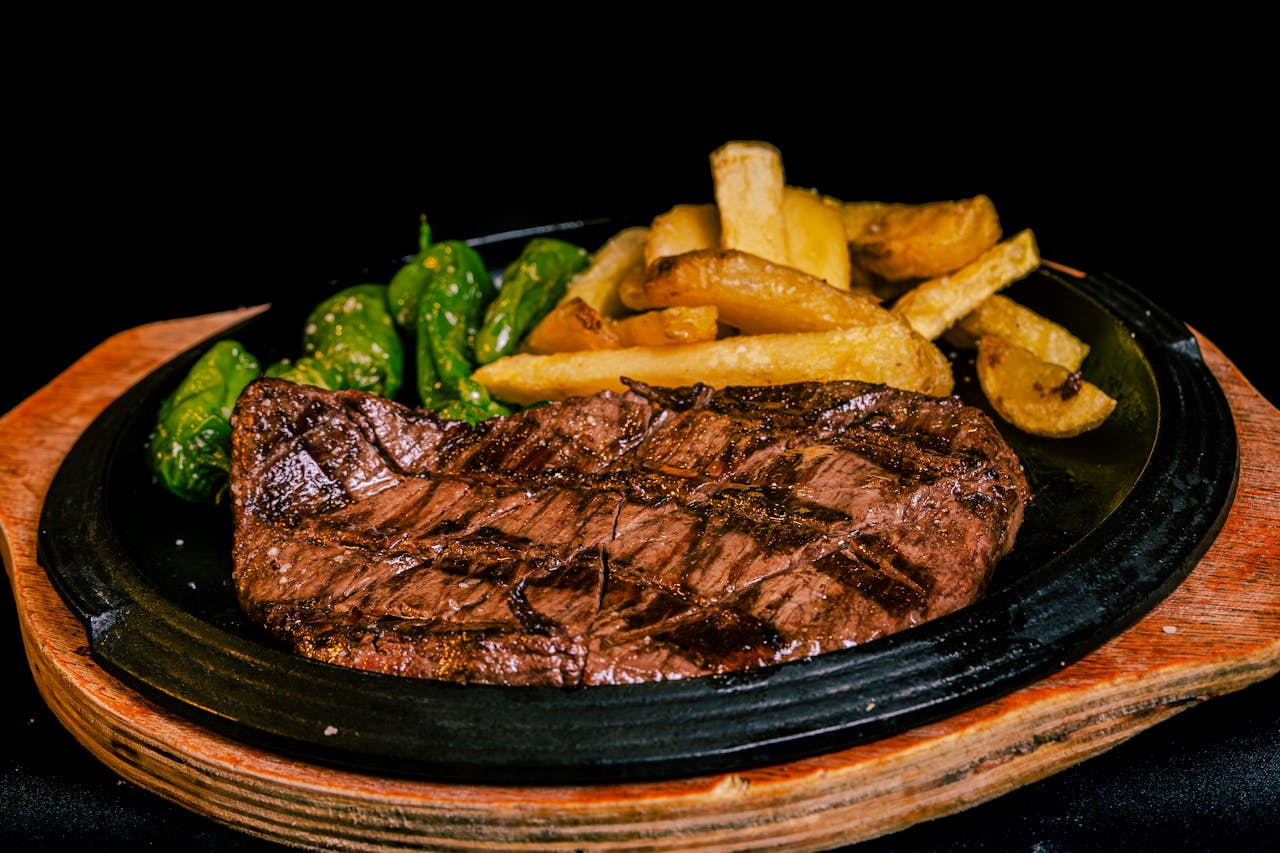 Grilled beef steak served with french fries and green peppers on a black plate.
